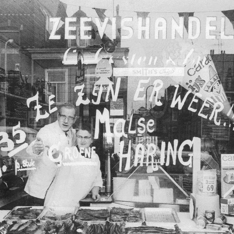 Zwart-witfoto uit het familiearchief: de etalage van de zeevishandel, met op de ruit geschilderd: Ze zijn er weer!! Malse haring. Achter het glas twee mensen in witte jassen.